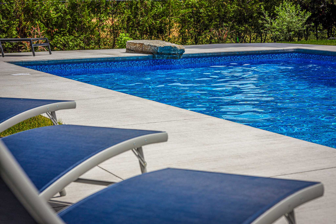 Modern inground swimming pool with a rich blue vinyl liner, surrounded by a smooth concrete deck. Two navy blue lounge chairs are positioned poolside, creating a relaxing atmosphere. A stone water feature gently spills into the pool, while a tall hedge provides privacy in the background.