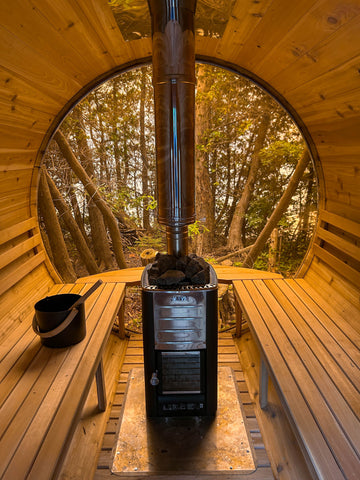 A black wood-burning stove in a red cedar sauna with a tall stainless steel chimney pipe, set inside a circular wooden sauna cabin with a view of a forest outside.