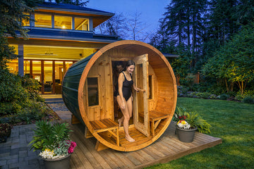 A woman in a black swimsuit stands in the doorway of a round wooden sauna cabin placed on a wooden deck in a backyard. The cabin has a dark green side panel and warm wood framing, with a house and trees visible in the background at dusk.