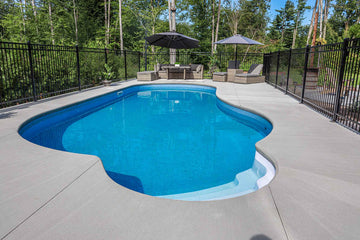 Curved vinyl inground swimming pool with integrated steps and spacious concrete deck, surrounded by a secure black metal fence and furnished with outdoor seating, installed by HB Pools in Navan, Ontario.