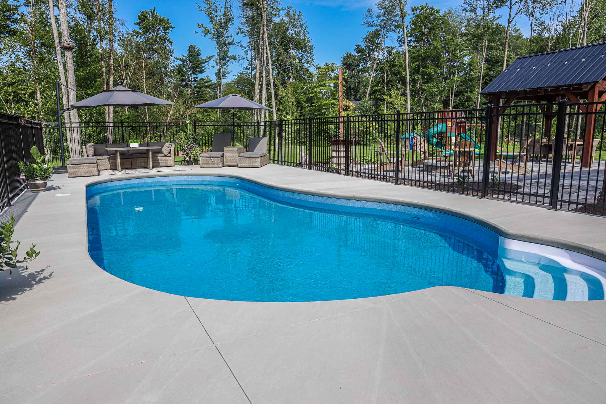 Custom vinyl inground swimming pool with curved steps and spacious concrete deck, installed by HB Pools in Navan, Ontario, featuring outdoor seating and a fenced backyard.