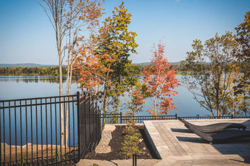 Scenic view of the Ottawa River & newly built pool area surrounded by autumn foliage, with vibrant orange and yellow leaves. The pool deck features modern pavers and lounge chairs, offering a relaxing spot to enjoy the tranquil waters and natural beauty. A black metal fence provides safety without obstructing the stunning view.