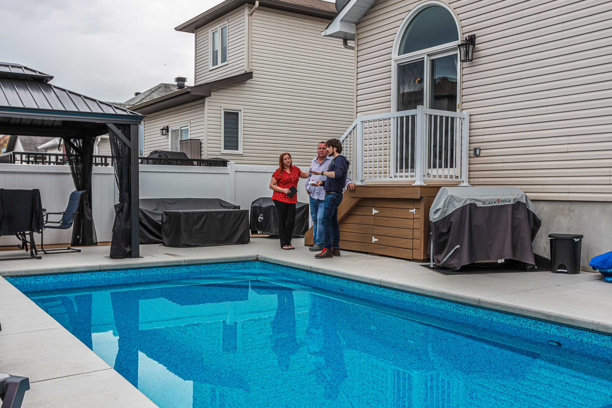 HB Pools technician consulting with homeowners beside a vinyl inground swimming pool in Rockland, Ontario, discussing pool features and maintenance in a residential backyard.