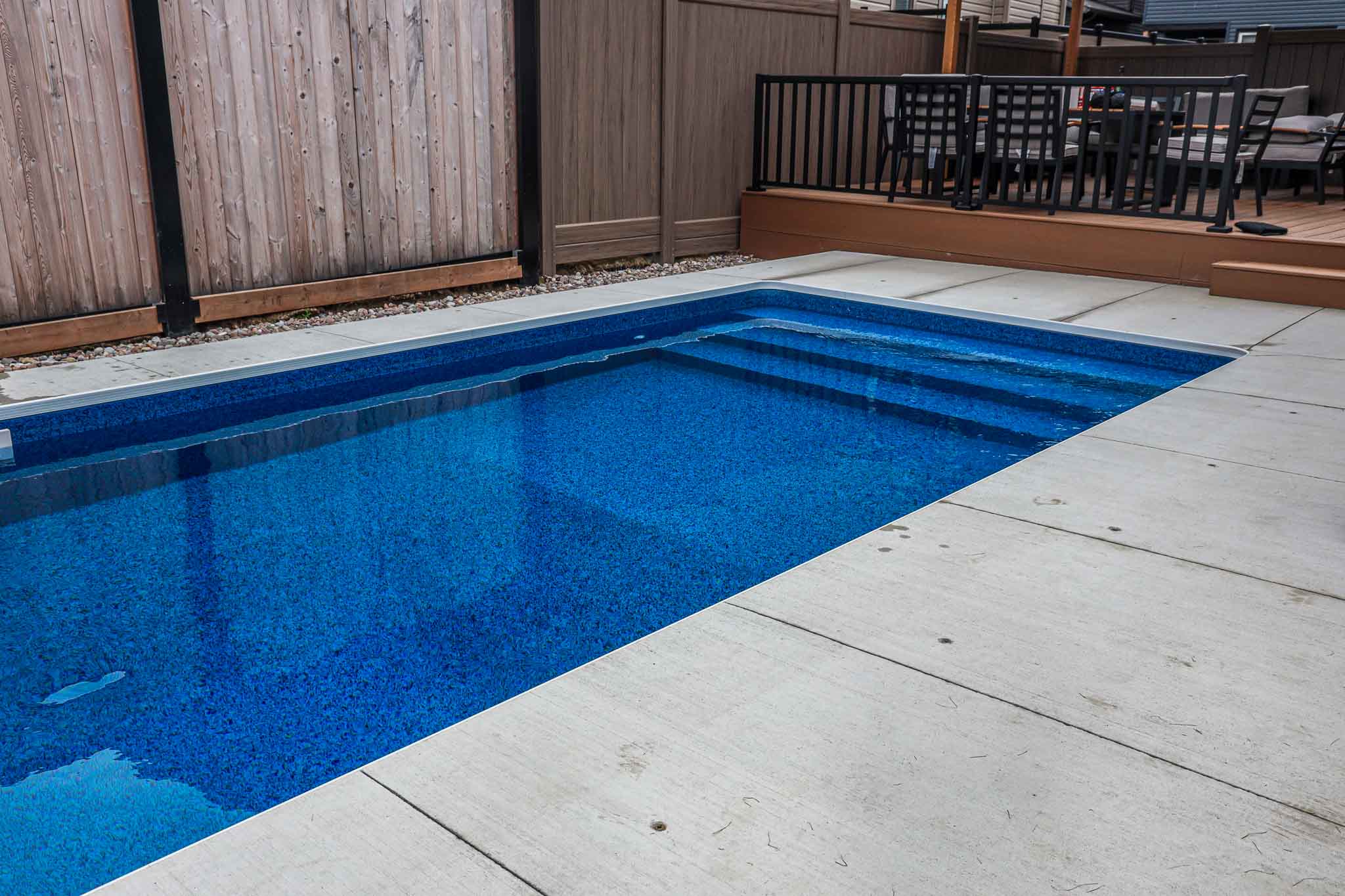 Rectangular vinyl inground swimming pool with built-in steps and surrounding concrete coping, adjacent to a raised wood deck, installed by HB Pools in Orleans, Ontario.