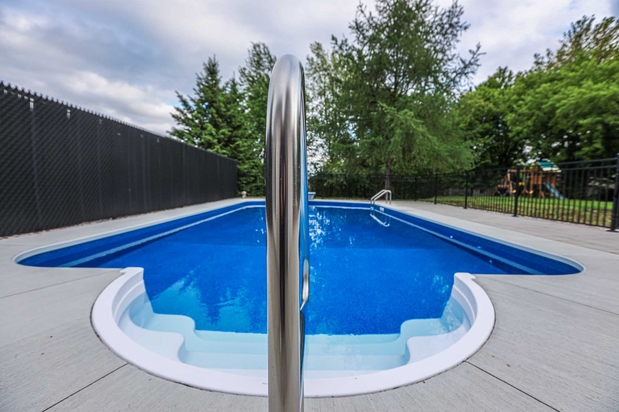 Vinyl inground swimming pool with curved steps and stainless steel handrail installed by HB Pools in Limoges, Ontario, surrounded by a clean concrete deck and privacy fencing.