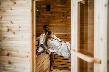 A couple relaxing in a wooden sauna, wearing white robes. The interior features light wood paneling and benches, with a glass door framing the scene.