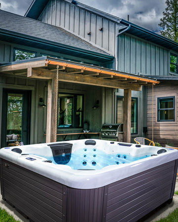 A large outdoor hot tub with a dark gray ribbed exterior and white interior, filled with blue water, situated on a patio in front of a house with gray siding and a wooden covered porch.
