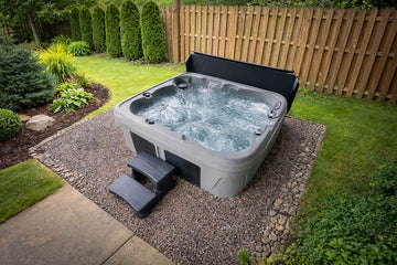A gray and black outdoor hot tub with bubbling water, set on a gravel pad in a landscaped backyard with grass, shrubs, and a wooden fence.