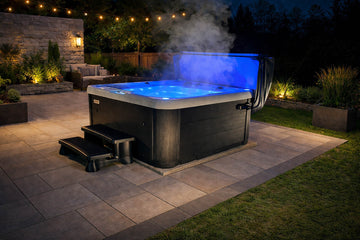 A hot tub with a dark gray exterior and a glowing blue interior, set on a stone patio in a backyard at night with steam rising from the water.