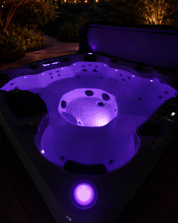 A hot tub at night with purple LED lighting illuminating the interior, photographed outdoors on a deck with garden foliage in the background.