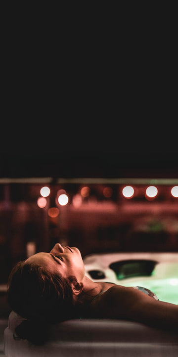 A woman relaxes in a spa hot tub at night, with warm string lights blurred in the background.