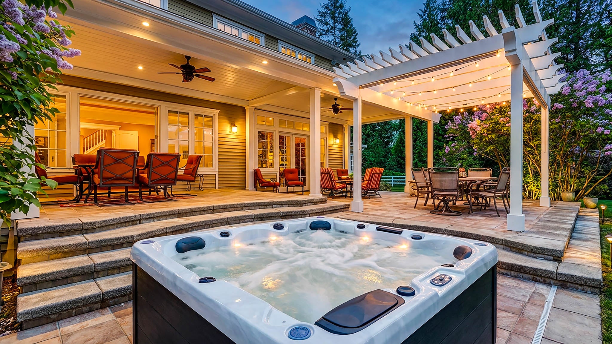 A hot tub with bubbling water in the foreground on a stone patio, with a house, outdoor dining area, pergola, and garden visible in the background at dusk.