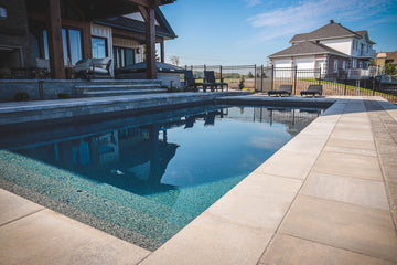 Modern backyard inground pool construction by HB Pools with crystal-clear water, surrounded by sleek pavers and adjacent to a stylish patio area with lounge seating. The pool area is enclosed by a black fence, providing both safety and elegance, while the nearby home and deck create a seamless flow between indoor and outdoor living