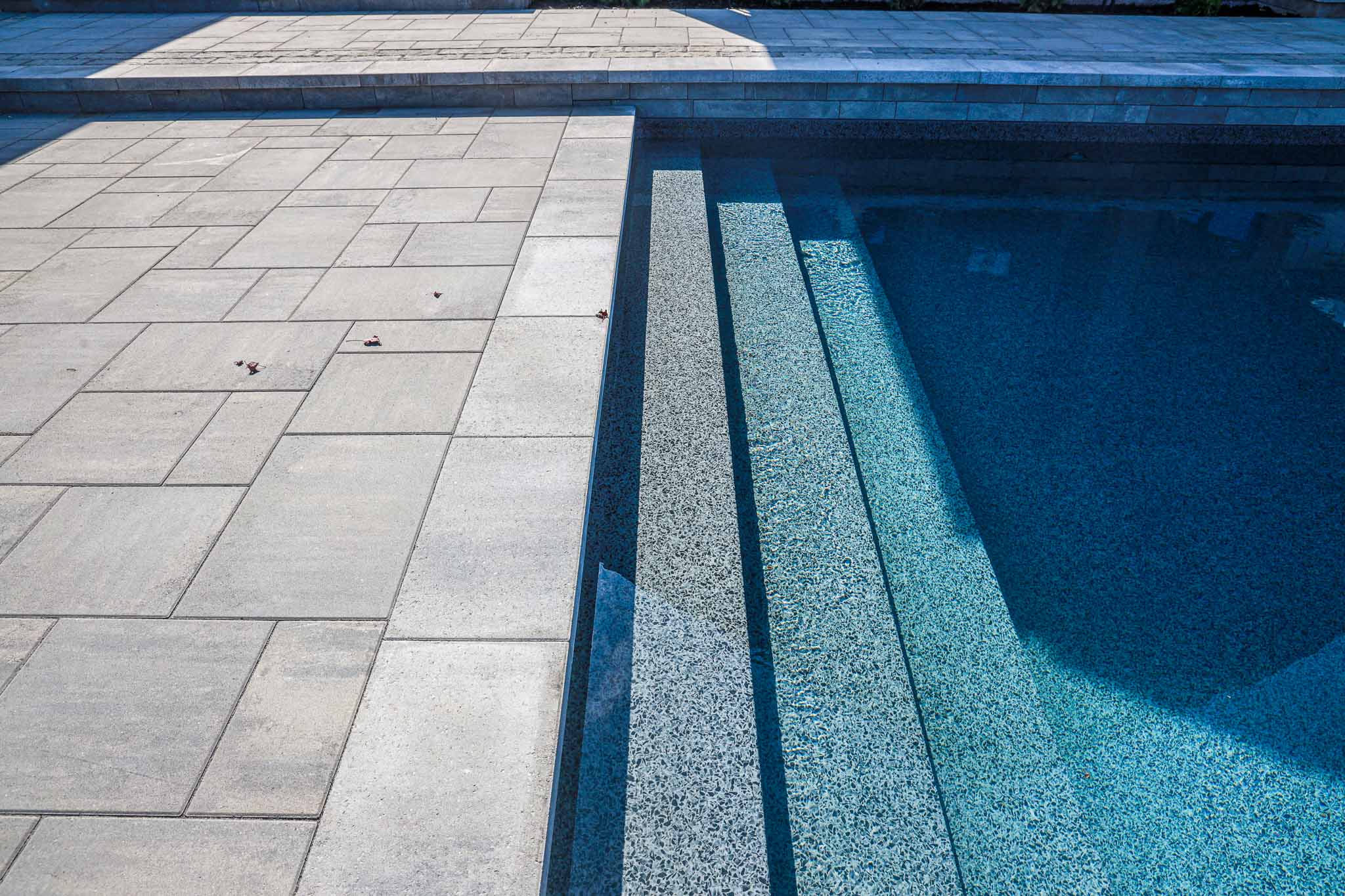 Close-up view of a custom pool's textured steps leading into clear blue water, surrounded by modern pavers. The design showcases a seamless transition between the pool area and the deck, emphasizing clean lines and a sleek, sophisticated look for a contemporary outdoor space