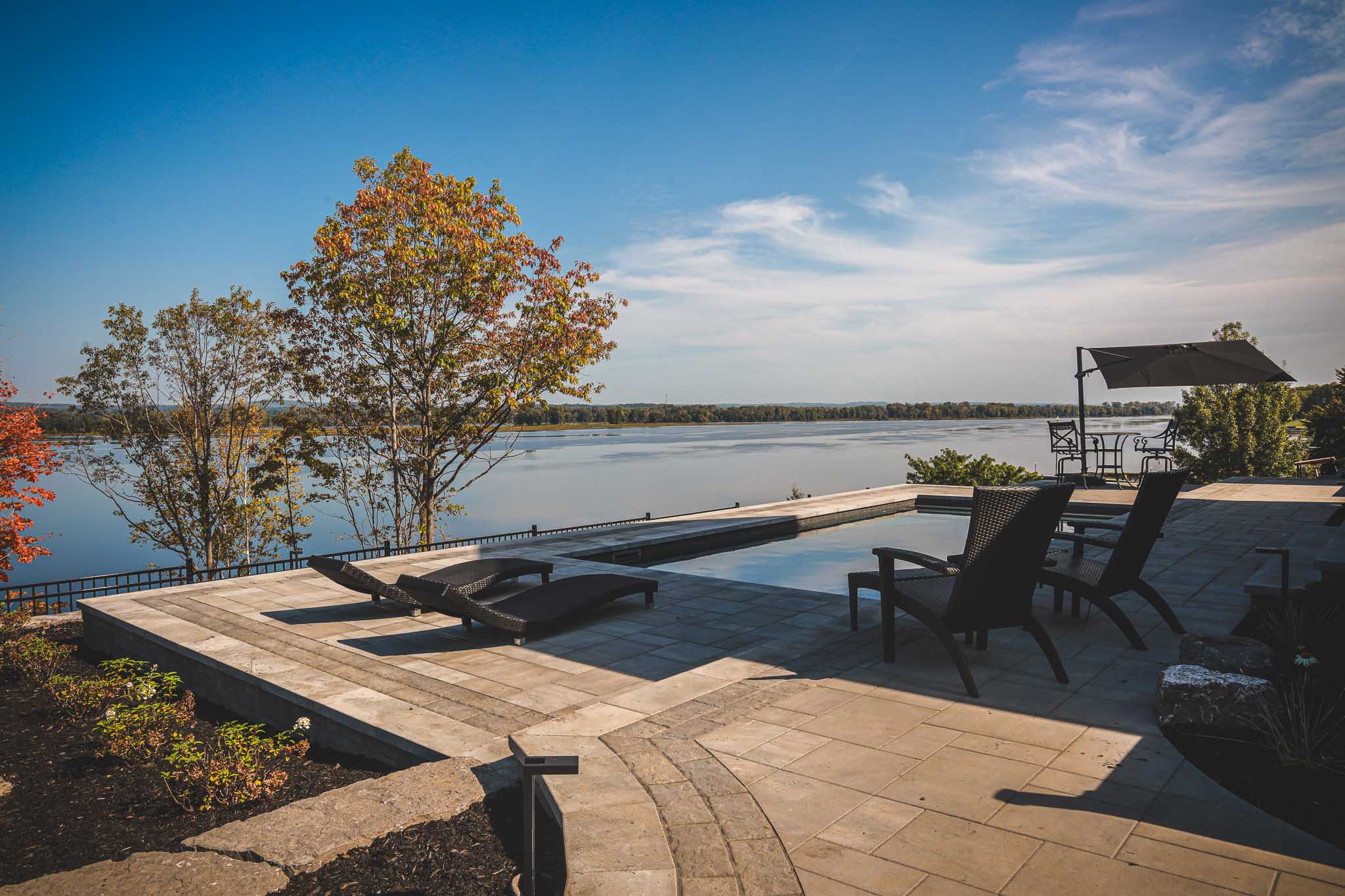 New Inground pool area with modern pavers, lounge chairs, and outdoor seating, surrounded by natural landscaping and trees with autumn foliage. The serene setting overlooks calm waters of the Ottawa River in Wendover, providing a tranquil atmosphere for relaxation and enjoying the beauty of nature. An umbrella offers shade over a table and chairs.