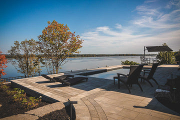 New Inground pool area with modern pavers, lounge chairs, and outdoor seating, surrounded by natural landscaping and trees with autumn foliage. The serene setting overlooks calm waters of the Ottawa River in Wendover, providing a tranquil atmosphere for relaxation and enjoying the beauty of nature. An umbrella offers shade over a table and chairs.