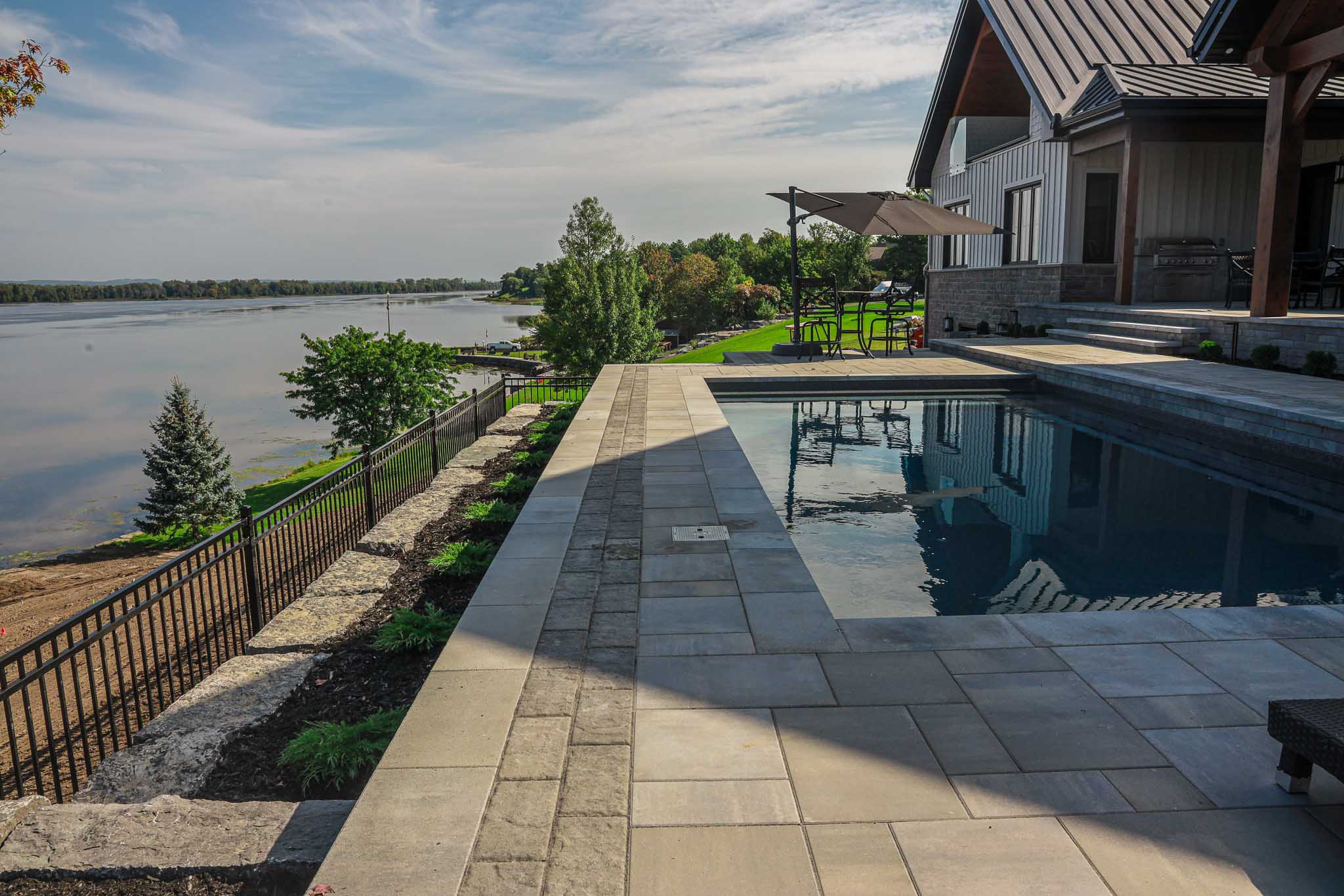 Elegant backyard pool area with a view of a the Ottawa River, featuring a modern pool surrounded by stylish pavers and landscaping. The poolside patio includes comfortable seating and an umbrella for shade, seamlessly integrating with the adjacent house and offering a perfect blend of relaxation and luxury