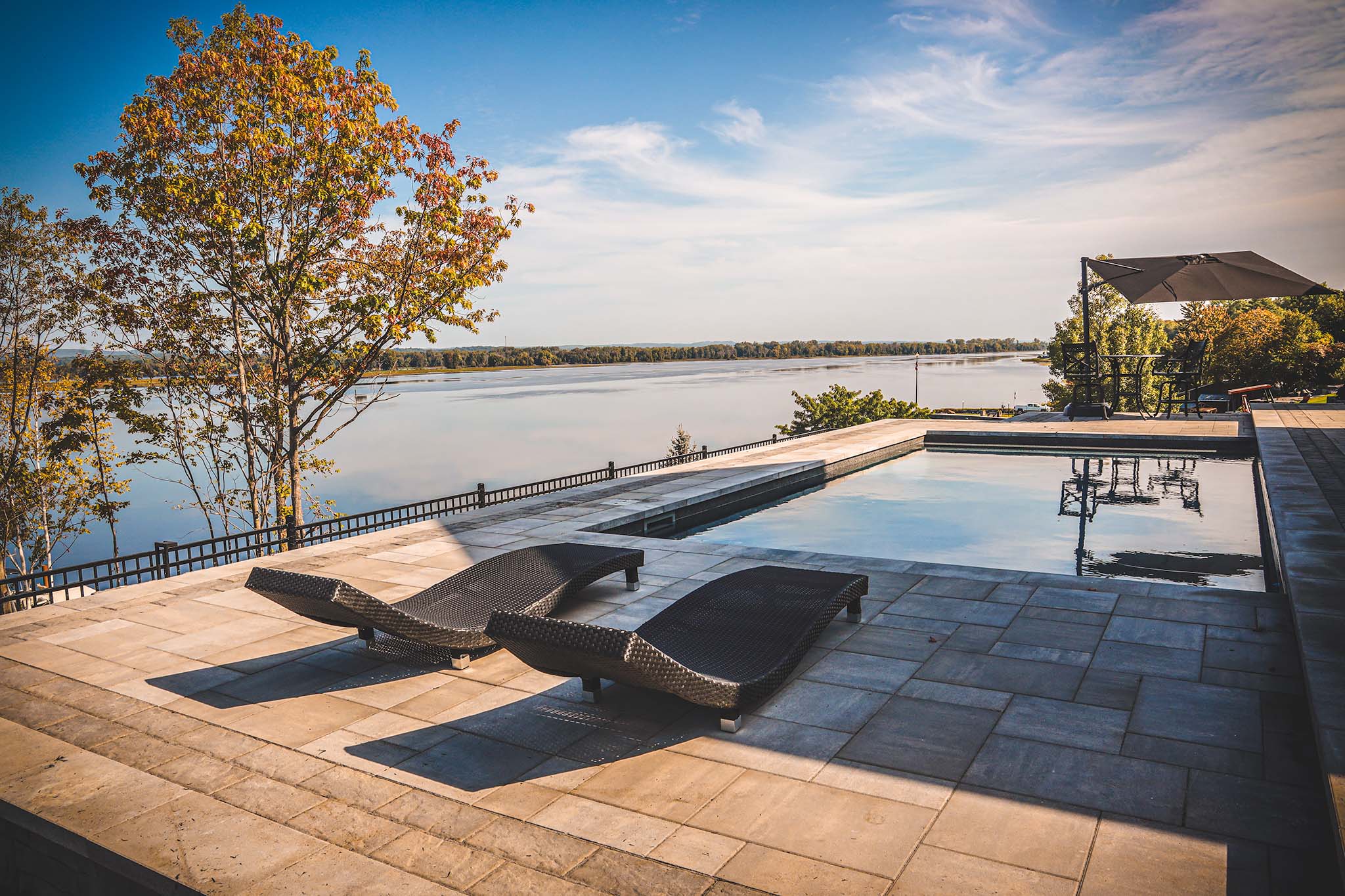 A luxurious Inground pool with sleek pavers overlooking the serene Ottawa River, surrounded by natural scenery with lush trees. The poolside features two modern lounge chairs positioned for relaxation, with reflections of the sky on the calm water. The setting exudes tranquility, perfect for enjoying the outdoors in Wendover Ontario.