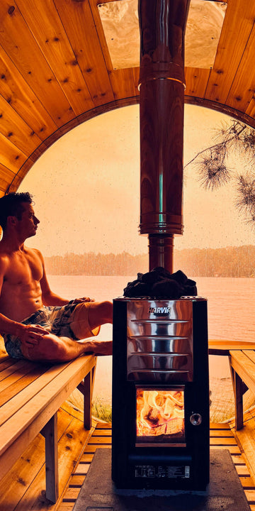 A man sits on a wooden bench inside a wooden sauna cabin structure next to a black wood-burning stove with a visible fire and tall metal chimney pipe. The background shows a warm-toned outdoor lake or river scene through an arched opening.